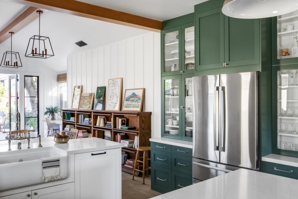 green cabinetry around a refrigerator and a long wooden bookcase displaying books and art.