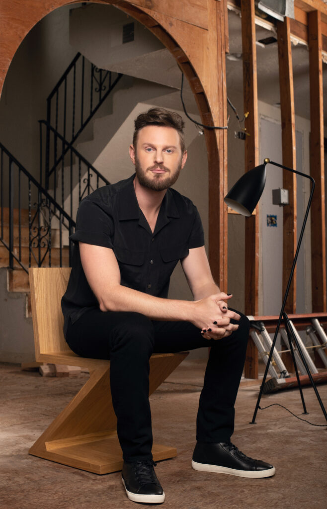 bobby berk wearing all black and sitting on a wooden chair