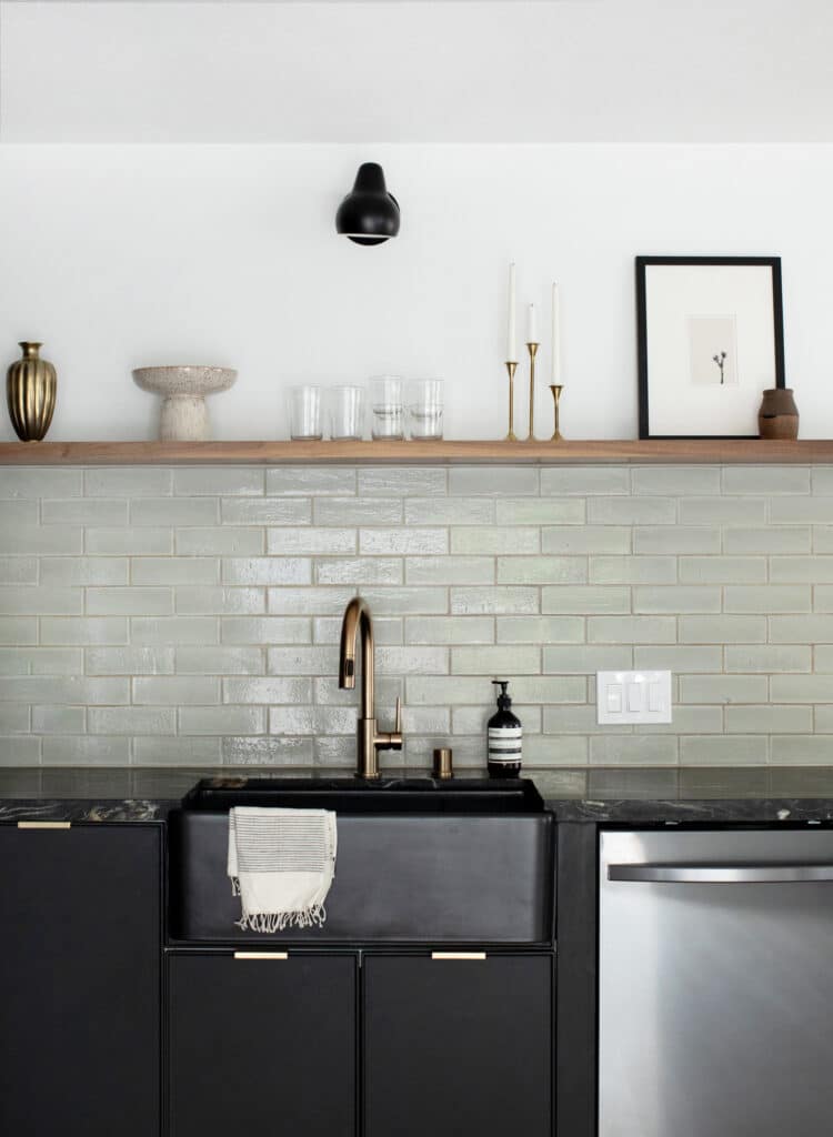 Black kitchen cabinets and sink with a sage green tile backsplash and open shelf